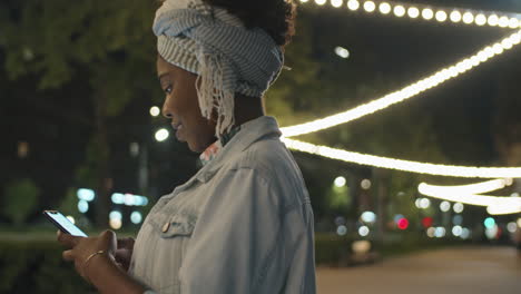 Mujer-Afroamericana-Enviando-Mensajes-Por-Teléfono-En-El-Parque-Por-La-Noche